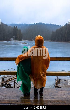 Zwei Kinder auf Regenmänteln genießen den mit Eis bedeckten See Synevyr, die Karpaten, Ukraine Stockfoto