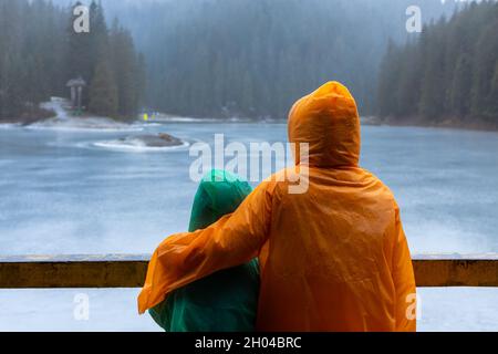 Zwei Kinder auf Regenmänteln genießen den mit Eis bedeckten See Synevyr, die Karpaten, Ukraine Stockfoto