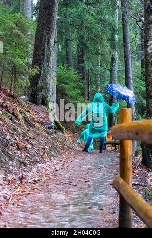 Frau mit Kindern auf Regenmänteln, die auf einem eisbedeckten Weg um den von Eis bedeckten See Synevyr, die Karpaten, Ukraine, wandern Stockfoto