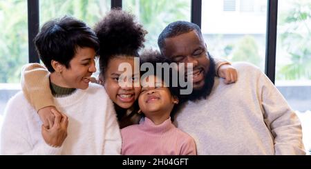 Afroamerikanischer Vater mit asiatischer Mischmutter zusammen mit zwei afrikanischen Töchtern im Pullover. Glückliche Familie kuschelt drinnen zusammen. Horizontale Panoramaansicht Stockfoto