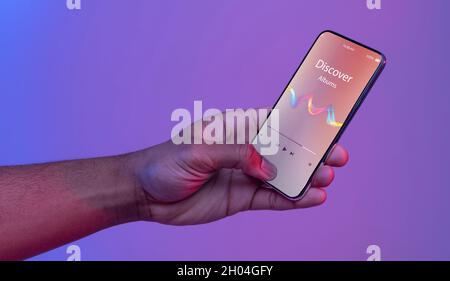 Schwarze männliche Hand hält Smartphone mit geöffneter Music Player App auf dem Bildschirm, während er unter Neonlicht über purpurem Hintergrund steht, unerkennbarer Mann E Stockfoto