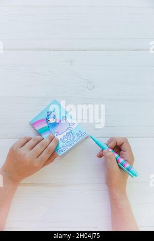 Hände eines Mädchens, das Notizbuch mit Lama-Einhorn und Regenbogen mit buntem Gelschreiber auf weißem Holzhintergrund hält. Idee des Girly Schreibtischs. Zurück zu sc Stockfoto