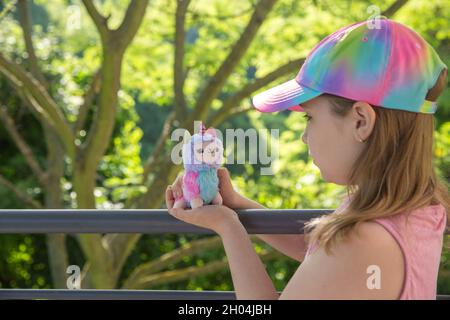 Farbige Plüsch Lama Einhorn in den Händen kleines Mädchen mit Grüns auf dem Hintergrund. Berühmtes und beliebtes Kinderspielzeug. Stockfoto