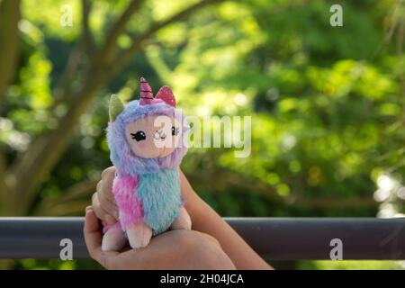 Farbige Plüsch Lama Einhorn in den Händen kleines Mädchen mit Grüns auf dem Hintergrund. Berühmtes und beliebtes Kinderspielzeug. Stockfoto