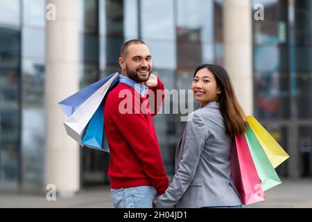 Verkaufssaison, riesige Rabatte Konzept. Zärtliches junges multirassisches Paar mit hellen Einkaufstaschen, die die Hände halten, die Kamera lächeln und zusammen gehen Stockfoto