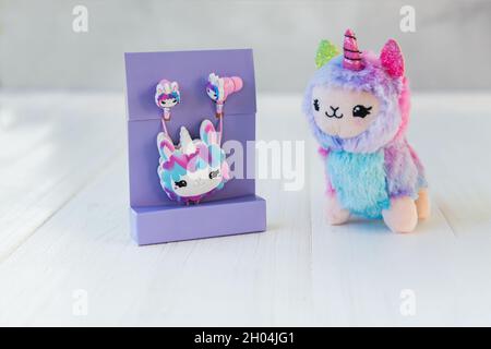 Verpackte Lama Einhorn Kopfhörer für Kinder in lila Packung und farbige Plüsch Lama Einhorn auf weißem Holz Hintergrund. Musik, Holiday Shopping Konzept. Stockfoto