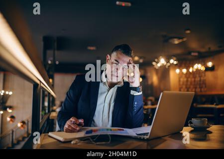 Ein besorgter Geschäftsmann mittleren Alters, der am Abend im Café saß und die Karte betrat und den Stift in der Hand hielt. Auf dem Tisch Kaffee, Laptop und Adend Stockfoto
