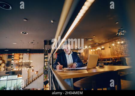 Lächelnder Geschäftsmann mittleren Alters, der am Abend im Café etwas auf die Tagesordnung gesetzt hat. Auf dem Tisch Kaffee und Laptop. Stockfoto