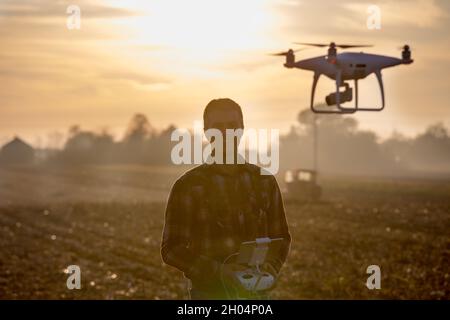 Attraktiver Landwirt, der Drohne über Ackerland mit Traktor im Hintergrund navigiert. Hightech-Innovationen zur Steigerung der Produktivität in der Landwirtschaft Stockfoto