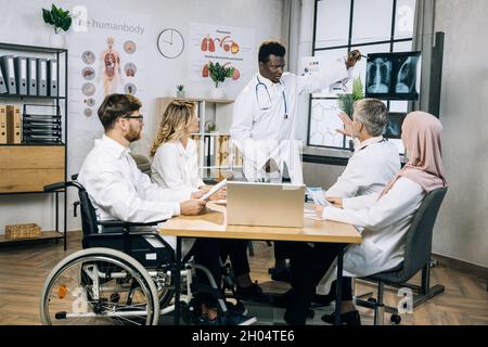 Gruppe von multirassischen medizinischen Spezialisten sitzen zusammen am Schreibtisch und hören afrikanische Kollegen, die auf Röntgenscan zeigen. Kompetente Ärzte prüfen die Ergebnisse der Patientenanalyse. Stockfoto