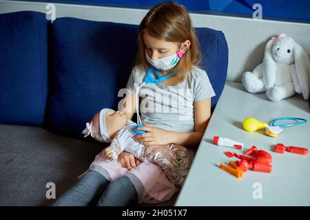 Ein kleines Mädchen in medizinischer Maske sitzt auf dem Sofa, spielt einen Arzt, behandelt und hört die Puppe mit einem Stethoskop in einer medizinischen Maske zu Hause während qu Stockfoto