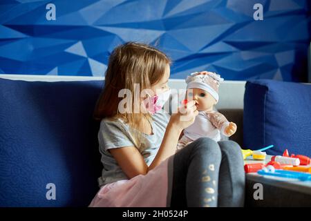 Ein kleines Mädchen in medizinischer Maske sitzt auf dem Sofa, spielt einen Arzt, behandelt und hört die Puppe mit einem Stethoskop in einer medizinischen Maske zu Hause während qu Stockfoto