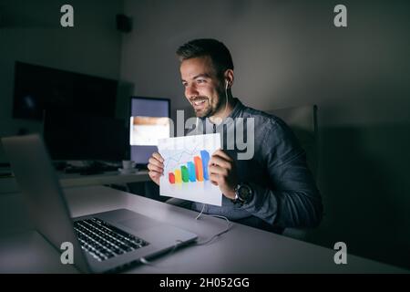 Junger lächelnder bärtiger Arbeiter, der in der späten Nacht im Büro saß und eine Telefonkonferenz und ein Diagramm vorstellte. Stockfoto