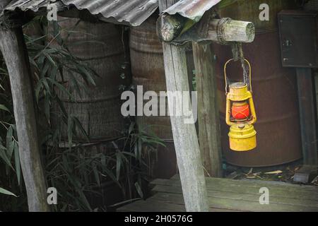 Alte schmuddelige gelbe Kerosin-Lampe mit rotem Glas hängt von einem Holzbalken unter einem schrägen Wellblechdach Stockfoto