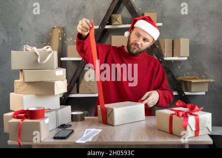 Ein Mann in einem Weihnachtsmütze bereitet in einem Lager Weihnachtsgeschenke zur Lieferung vor und bindet ein rotes Band ein. Kartons auf dem Tisch. Neujahrsangebot Stockfoto