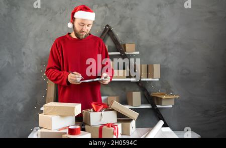 Kaukasischen bärtigen Mann trägt weihnachtsmann Hut und roten Pullover Business-Besitzer zu Hause arbeiten. weihnachtsgeschenk Einkaufen concept.Marks Boxen für die Lieferung Stockfoto
