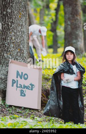 Vertikales Foto eines ernsthaften Freiwilligen-Mädchens, das in der Nähe des Pappkartons an einem Baum mit der Aufschrift No Planet B und einem Mann im Hintergrund steht Stockfoto