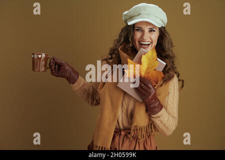 Hallo oktober. Glückliche moderne 40 Jahre alte Hausfrau in Schal mit gelben Herbst Ahornblatt, Buch und Lederhandschuhe isoliert auf beigem Hintergrund. Stockfoto
