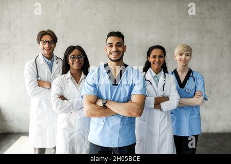 Internationales Ärzteteam, das beim Morgenbreefing zusammen posiert, medizinische Angestellte und Angestellte, die gegen die Wand stehen und die Arme mit dem Kreuz halten Stockfoto