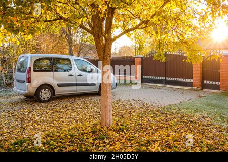 Silberner Kleinbus, der an einem sonnigen Herbsttag auf einem Hof geparkt wurde. Das goldene Laub fällt vom Birnenbaum im Licht der Sonnenstrahlen. Ein Haufen gelb gefallener Blätter auf dem Bürgersteig. Stockfoto