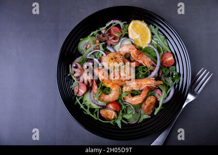 Salat mit Meeresfrüchten, Rucola, Tomaten, Gurken, roter Zwiebel und Zitrone auf einem schwarzen Teller. Schwarzer Hintergrund, Nahaufnahme. Stockfoto