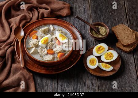 Zuek polieren fermentierte Roggensuppe mit traditioneller polnischer weißer Kielbasa oder Wurst mit Majoran, hart gekochte Eier, die auf einer Tonschüssel mit einer Holzschale serviert werden Stockfoto