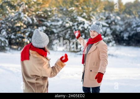 Fröhlicher Kerl in warmen Outfit mit Schnee kämpfen mit seiner Freundin, glücklich liebende Paar genießen sonnigen Wintertag im Wald, warf Schneebälle auf jeden Stockfoto