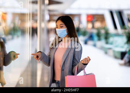 Junge asiatische Frau in Gesichtsmaske hält Geschenktüten, Einkaufen für neue Kleidung in riesigen Supermarkt. Millennial Dame beim Kauf von Waren im Einkaufszentrum während covid-19 l Stockfoto