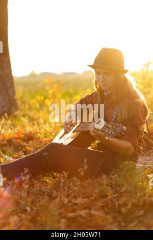 Unschärfe junge Frau spielen Gitarre bei Sonnenuntergang im Herbst Feld. Offene Silhouette Frau in Hut chill spielen akustische oder klassische Gitarre Musiker. Aufflackern der Sonne Stockfoto