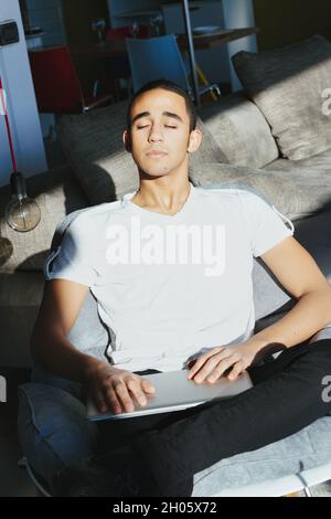 Der junge Mann genießt die warme Sommersonne, während er auf einem Sofa sitzt, mit geschlossenen Augen und glückseligem Ausdruck, während er an seinem Laptop arbeitet Stockfoto
