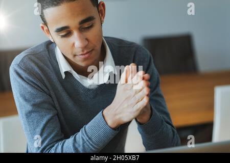 Der junge schwarze Geschäftsmann in einem Großraumbüro blickt pensiver auf seinen Computer mit zusammengeschnittenen Händen in einem Nahaufnahme-Porträt Stockfoto