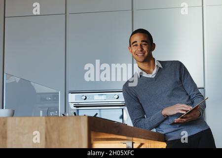 Fröhlicher, freundlicher, junger schwarzer Geschäftsmann, der sich auf eine Küchentheke stützt und seinen Tablet-pc mit einem strahlenden Lächeln auf die Kamera grinsend hält Stockfoto