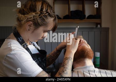 SANTIAGO, CHILE - 27. Dez 2018: Eine Barbierin nimmt bei einem traditionellen Barbier in San einen Rasiermesser an den Bart ihres rothaarigen männlichen Kunden Stockfoto