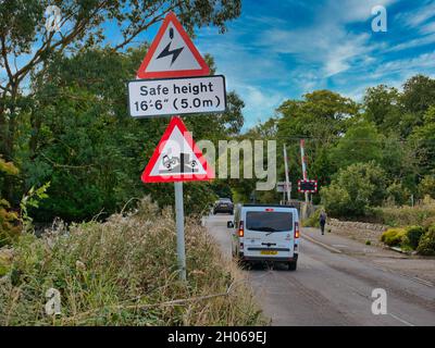 Fahrzeuge passieren Schilder, die vor elektrischen Kabeln über dem Kopf und einer Gefahr der Erdung warnen, wenn sie sich einem Bahnübergang in Beal, Northumberland, Großbritannien, nähern. Stockfoto