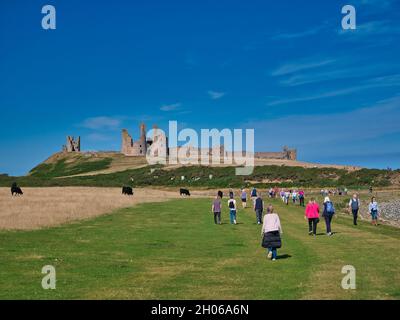 Touristen nähern sich den Ruinen des Dunstanburgh Castle aus dem 14. Jahrhundert an der Küste von Northumberland im Nordosten Großbritanniens. An einem sonnigen Tag aufgenommen. Stockfoto