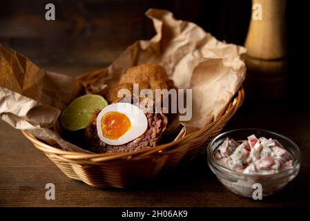 Portion Scotch-Ei und Pico de gallo-Sauce auf сraft-Papier und Korbkorb. Gebratenes Pub-Essen im Inneren einer Bar Stockfoto