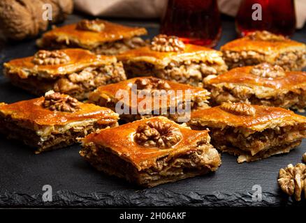 Honig Baklava mit Walnüssen auf schwarzem Schiefer. Portionen orientalischer Süßigkeiten. Türkische zuckerhaltige Stücke. Im Hintergrund gegossene Tees in Schmuduglas. Stockfoto