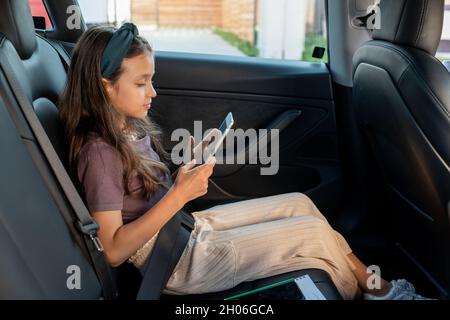 Seitenansicht des niedlichen Mädchen Scrollen im Smartphone, während auf dem Rücksitz des Elektroautos sitzen Stockfoto