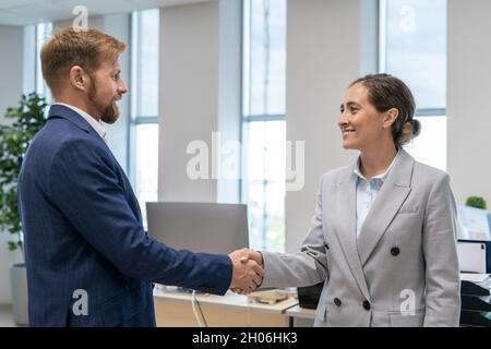 Zwei glückliche Geschäftspartner in eleganten Anzügen, die im Büro die Hände gegen den Arbeitsplatz schütteln Stockfoto
