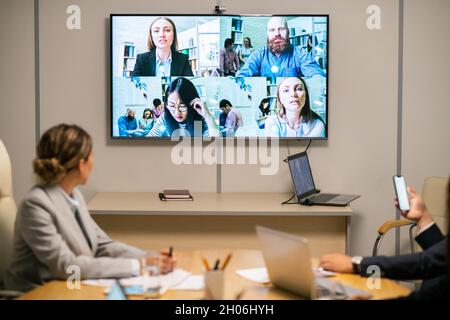 Mehrere Geschäftsleute auf einem großen Display, das an der Wand des Sitzungssitzes hing, wo eine Gruppe von Mitarbeitern ein Webinar abgehalten hat Stockfoto