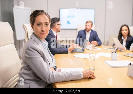 Blonde hübsche Geschäftsfrau in grauer formalwear sitzt am Tisch gegen eine Gruppe von Mitarbeitern Stockfoto