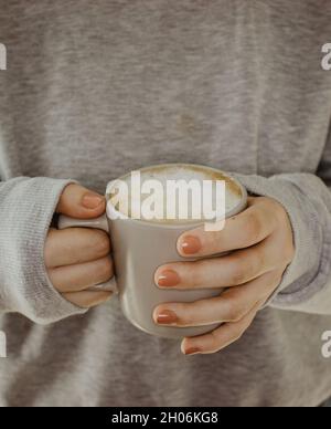 Nahaufnahme der weiblichen Hände mit einer Tasse Getränke. Schöne graue Pullover Mädchen hält eine Tasse Tee oder Kaffee in der Morgensonne. Becher für Ihr Design. Stockfoto