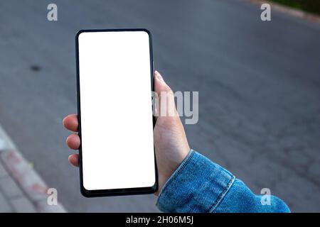 Frau mit schwarzem Smartphone. Mockup-Bild einer Frau, die das Mobiltelefon mit einem leeren weißen Desktop-Bildschirm hält, während sie auf dem Bürgersteig läuft. Stockfoto