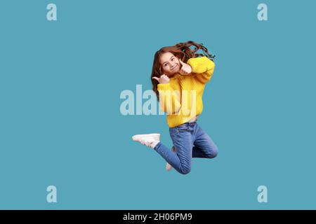 In voller Länge Porträt von glücklich charmant kleines Mädchen springen hoch und zeigt Daumen bis zur Kamera, tragen gelben lässigen Stil Pullover. Innenaufnahme des Studios isoliert auf blauem Hintergrund. Stockfoto