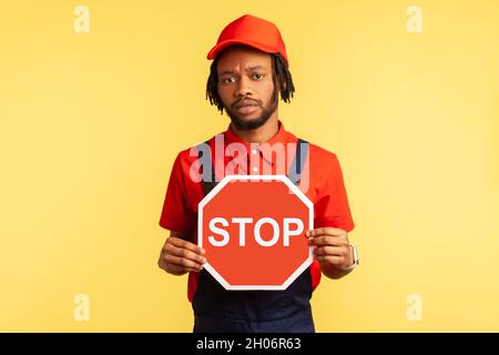 Porträt eines Handwerkers mit blauen Overalls, rotem Hut und T-Shirt mit rotem Stoppschild, Verbote, ernsthafter Blick auf die Kamera. Innenaufnahme des Studios isoliert auf gelbem Hintergrund. Stockfoto