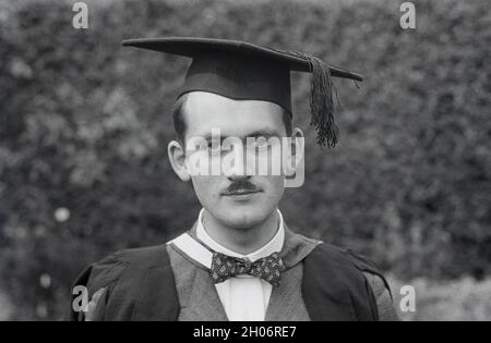 Historisches Nahaufnahme-Bild aus den 1950er Jahren eines jungen Mannes mit einem kurzen Schnurrbart und einem zweireihigen Anzug und einer Fliege, der für sein Bild in einem Abschlusskleid und einer quadratischen akademischen Mütze posiert, oft als Mütze und Kleid bezeichnet, England, Großbritannien. Seine spezielle Art von Mütze wird bei Abschlussfeiern mit akademischen Regalia getragen und wird als Mortarboard oder manchmal auch als Oxford Cap bezeichnet. Sie besteht aus einem horizontalen quadratischen Brett, das auf einer Schädelkappe mit einer Quaste in der Mitte befestigt ist. Sein unverwechselbares Aussehen bedeutet, dass die Obergrenze mit der akademischen Welt in Verbindung gebracht wird. Stockfoto