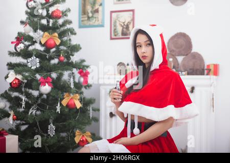 Junge chinesische Frau, die als weihnachtsmann gekleidet ist und eine elektronische Zigarette in der Nähe des weihnachtsbaums hält Stockfoto