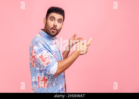 Seitenansicht eines jungen erwachsenen Mannes, der mit schockiertem, staunenden Gesichtsausdruck auf das Mobiltelefon zeigt, mit großen Augen und offenem Mund auf die Kamera blickt, Aufnahme des Innenstudios isoliert auf rosa Hintergrund. Stockfoto