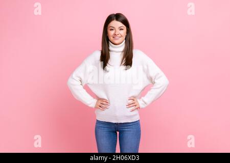 Optimistisches Mädchen mit dunklem Haar, das mit den Händen auf den Hüften steht, die Kamera anschaut, Selbstvertrauen ausdrückt und einen weißen Pullover im lässigen Stil trägt. Innenaufnahme des Studios isoliert auf rosa Hintergrund. Stockfoto