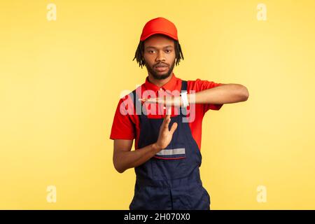 Seriöse Arbeiter tragen Uniform, zeigen Auszeit-Geste, brauchen Pause, arbeiten in der Dienstleistungsbranche, Kurierdienst, Housekeeping Wartung. Innenaufnahme des Studios isoliert auf gelbem Hintergrund. Stockfoto